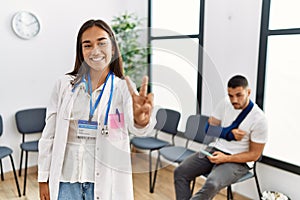 Young asian doctor woman at waiting room with a man with a broken arm smiling looking to the camera showing fingers doing victory