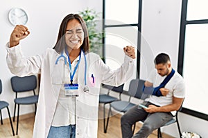 Young asian doctor woman at waiting room with a man with a broken arm very happy and excited doing winner gesture with arms