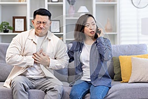 Young Asian family sitting on sofa at home. A man holds his hand to his chest, heart attack, stroke, panic attack, feels