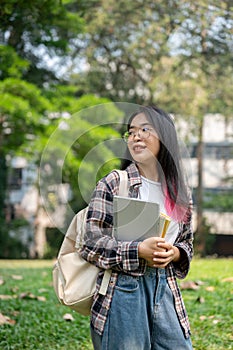 A young Asian female college student carrying a backpack and stuff, standing in the campus park