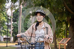 A young Asian female college student wearing a bike helmet is pushing her bicycle in a green park