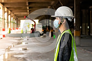 Young Asian female construction engineer wearing a hat and a green safety vest, stands looking at work in the construction zone