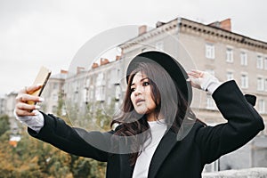 Young asian female in dark coat and hat taking a selfie outdoors