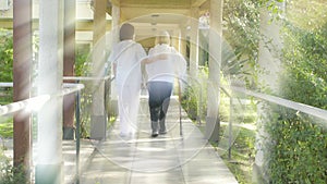 Young Asian female doctor assisting elderly woman with crutches in hospital garden. Back view. Light rays from behind