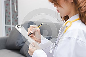 Young Asian female doctor writing notes on clipboard in hospital office.