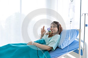 Young Asian female patient is smiling and showing ok gesture on hospital bed in hospital room. Medical healthcare concept
