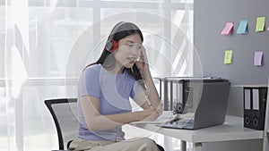 Young Asian female student wearing headphones surfs the internet on a laptop while taking an online