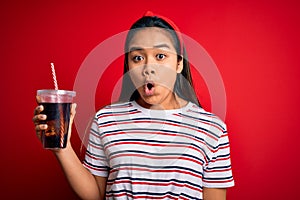 Young asian girl drinking cola fizzy refreshment using straw over isolated red background scared in shock with a surprise face,