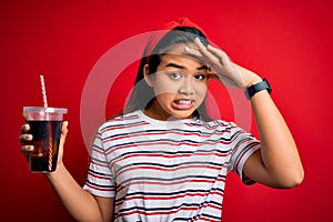 Young asian girl drinking cola fizzy refreshment using straw over isolated red background stressed with hand on head, shocked with