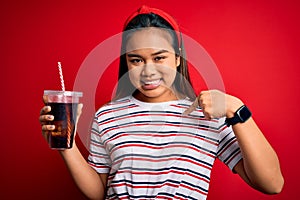 Young asian girl drinking cola fizzy refreshment using straw over isolated red background with surprise face pointing finger to