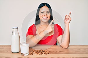 Young asian girl drinking healthy almond milk smiling and looking at the camera pointing with two hands and fingers to the side