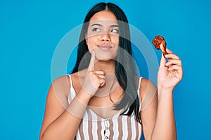 Young asian girl eating chicken wings serious face thinking about question with hand on chin, thoughtful about confusing idea