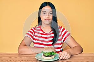 Young asian girl eating a tasty classic burger thinking attitude and sober expression looking self confident