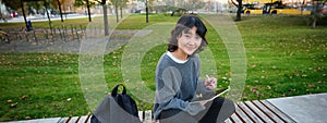 Young asian girl with graphic pencil and tablet, sits in park on bench, draws scatches, does her homework outdoors
