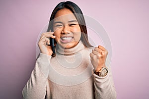 Young asian girl having conversation talking on the smartphone over isolated pink background screaming proud and celebrating