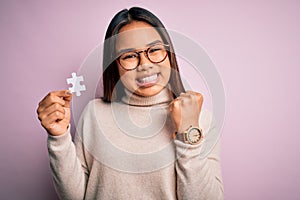 Young asian girl holding piece of puzzle standing over isolated pink background screaming proud and celebrating victory and