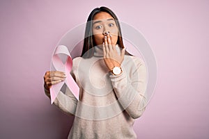Young asian girl holding pink cancer ribbon symbol for suppport over isolated background cover mouth with hand shocked with shame