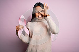 Young asian girl holding pink cancer ribbon symbol for suppport over isolated background with happy face smiling doing ok sign