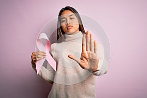 Young asian girl holding pink cancer ribbon symbol for suppport over isolated background with open hand doing stop sign with