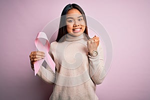 Young asian girl holding pink cancer ribbon symbol for suppport over isolated background screaming proud and celebrating victory