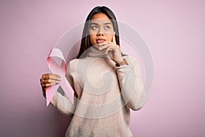 Young asian girl holding pink cancer ribbon symbol for suppport over isolated background serious face thinking about question,