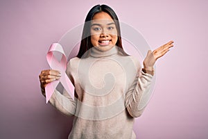 Young asian girl holding pink cancer ribbon symbol for suppport over isolated background very happy and excited, winner expression