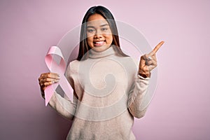 Young asian girl holding pink cancer ribbon symbol for suppport over isolated background very happy pointing with hand and finger