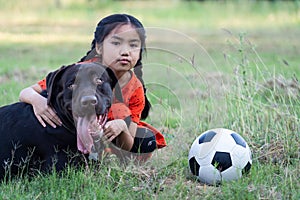 A young Asian girl playing football with her big black dog outside the grass ground in the yard in the evening