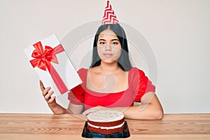 Young asian girl wearing birthday hat holding present thinking attitude and sober expression looking self confident