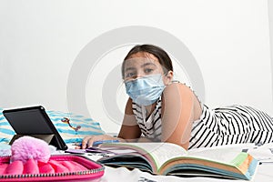 Young Asian girl wearing protective face mask laying on her bed studying.