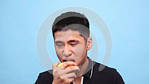 Young asian guy biting lemon against blue studio background. Handsome young man with grimacing face eating sour citrus