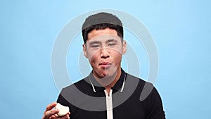 Young asian guy biting onio against blue studio background. Handsome young man with grimacing face eating spicy, bitter