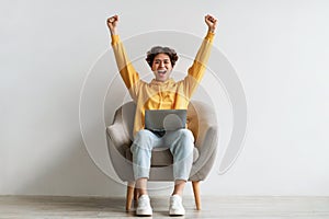 Young Asian guy sitting in armchair with laptop, raising hands up in excitement, celebrating success against white wall