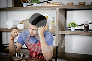 Young Asian handsome man feels stressed and bored, touched head while eating, having breakfast meal on wooden table in kitchen