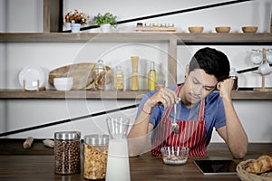 Young Asian handsome man feels stressed and bored, touched head while eating, having breakfast meal on wooden table in kitchen