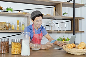 Young Asian handsome man is having, eating cereals and cornflakes in breakfast meal while using smart phone and laptop for working