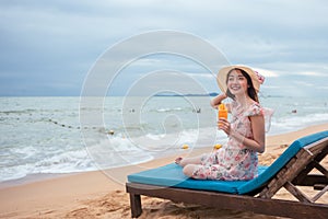 Asian happy woman wearing sun hat holding orange juice relaxing on chair at the beach with happily