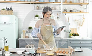 Young Asian housewife in kitchen using tablet for searching recipes online cooking the bakery dough homemade