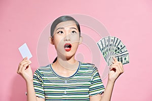 Young asian lady holding credit card and fan of money, posing isolated over pink studio background, girl doing shopping online