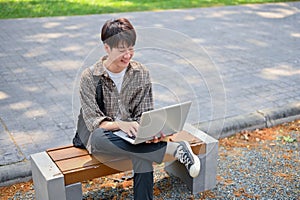 A young Asian male college student using his laptop computer on a bench in the campus park