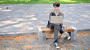 A young Asian male college student using his laptop computer on a bench in the campus park