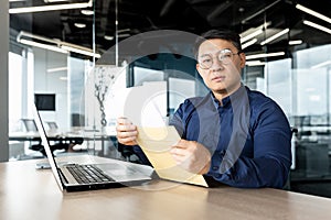 Young Asian male lawyer in glasses sitting in the office at a desk with a laptop. He holds an envelope with documents in