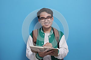 Young Asian male student wearing green baseball jacket writing on book