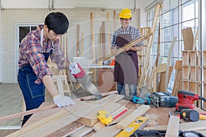 Young asian male worker wearing plaid shirt and blue jean apron with white gloves holding saw cutting wood stick on the table full