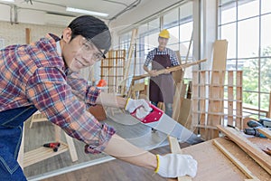 Young asian male worker wearing plaid shirt and blue jean apron with white gloves holding saw cutting wood stick on the table full