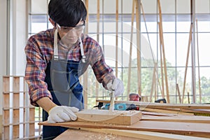 Young asian male worker wearing plaid shirt and blue jean apron with white gloves holding saw cutting wood stick on the table full