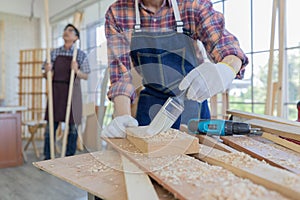 Young asian male worker wearing plaid shirt and blue jean apron with white gloves holding saw cutting wood stick on the table full