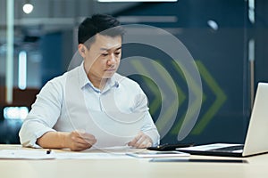 Young Asian male working with documents and calculator. Sitting with a laptop in a modern office