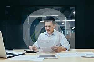 Young Asian male working with documents and calculator. Sitting with a laptop in a modern office