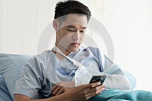 Young asian man with arm in sling lying on hospital bed looking at mobile phone. Broken arm patient